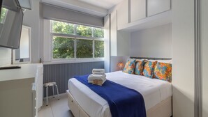 Quarto com cama, janela, radiador, um banquinho pequeno e televisão.