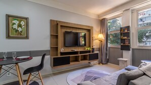 Uma sala moderna com televisão de tela plana, área de jantar com cadeiras, um sofá e uma janela com vista para edifícios.