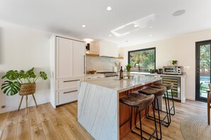 A modern kitchen with a marble island, bar stools, and a view of greenery outside.