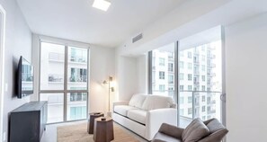 A modern apartment with a white sofa, a TV on the wall, and large windows offering a view of other buildings. - Downtown Condo with Pool, Gym & City Views (Miami)