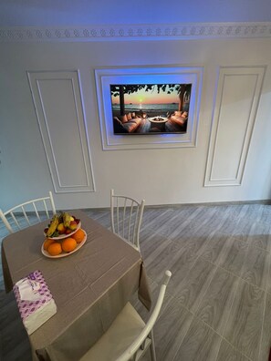 A dining area with a table set for two, a fruit bowl, and a television mounted on the wall displaying a sunset scene. - Cozy Apartment Netflix WiFi Food Pickup In Tangier (Tangier)