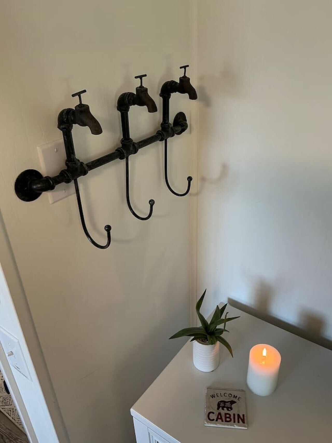 Un sistema di ganci neri fissato al muro, con una candela accesa e un cartello di benvenuto su un tavolo bianco.