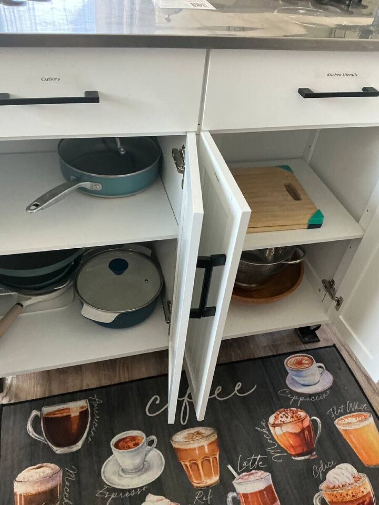 Una cocina con un cajón etiquetado 'Cutlery', un estante con una olla azul y una pizarra con ilustraciones de bebidas de café.