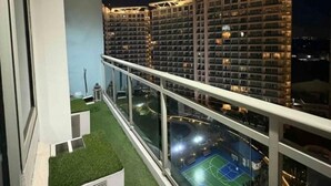 Un balcon donnant sur un court de tennis et sur les immeubles de grande hauteur situés à proximité, le tout illuminé la nuit.