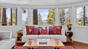 A modern living room with a white sofa, red and gold cushions, a wooden coffee table, and large windows offering a view of trees and a body of water. - Kings Beach Cabin w/ Panoramic Lakeview & Forest (Kings Beach)