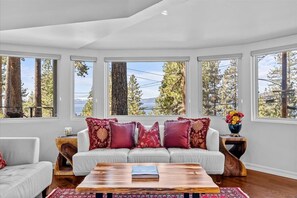 A modern living room with a white sofa, red and gold cushions, a wooden coffee table, and large windows offering a view of trees and a body of water. - Kings Beach Cabin w/ Panoramic Lakeview & Forest (Kings Beach)