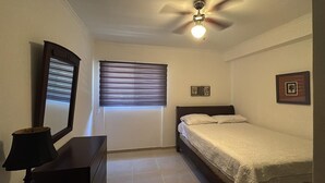 A bedroom with a bed, a ceiling fan, a window with blinds, a dresser, and a framed picture on the wall.