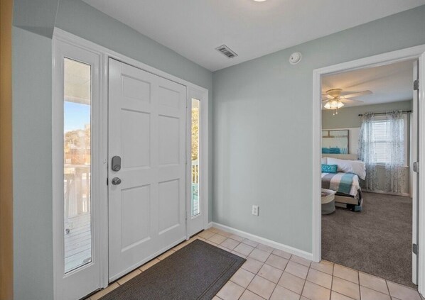 A hallway with a white door, a doormat, and a view into a bedroom with a bed, curtains, and a ceiling fan.