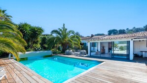 Une piscine dotée d’une terrasse en bois, entourée de palmiers et bordée d’une maison moderne aux grandes fenêtres.