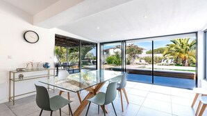 Un espace repas moderne avec une table en verre, des chaises, une horloge et une vue sur une piscine et un jardin.