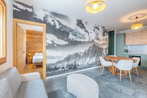 Ein modernes Wohnzimmer mit Wandgemälde von Bergen, ein Essbereich mit Holztisch und -stühlen sowie eine Küche mit hellgrünen Schränken.