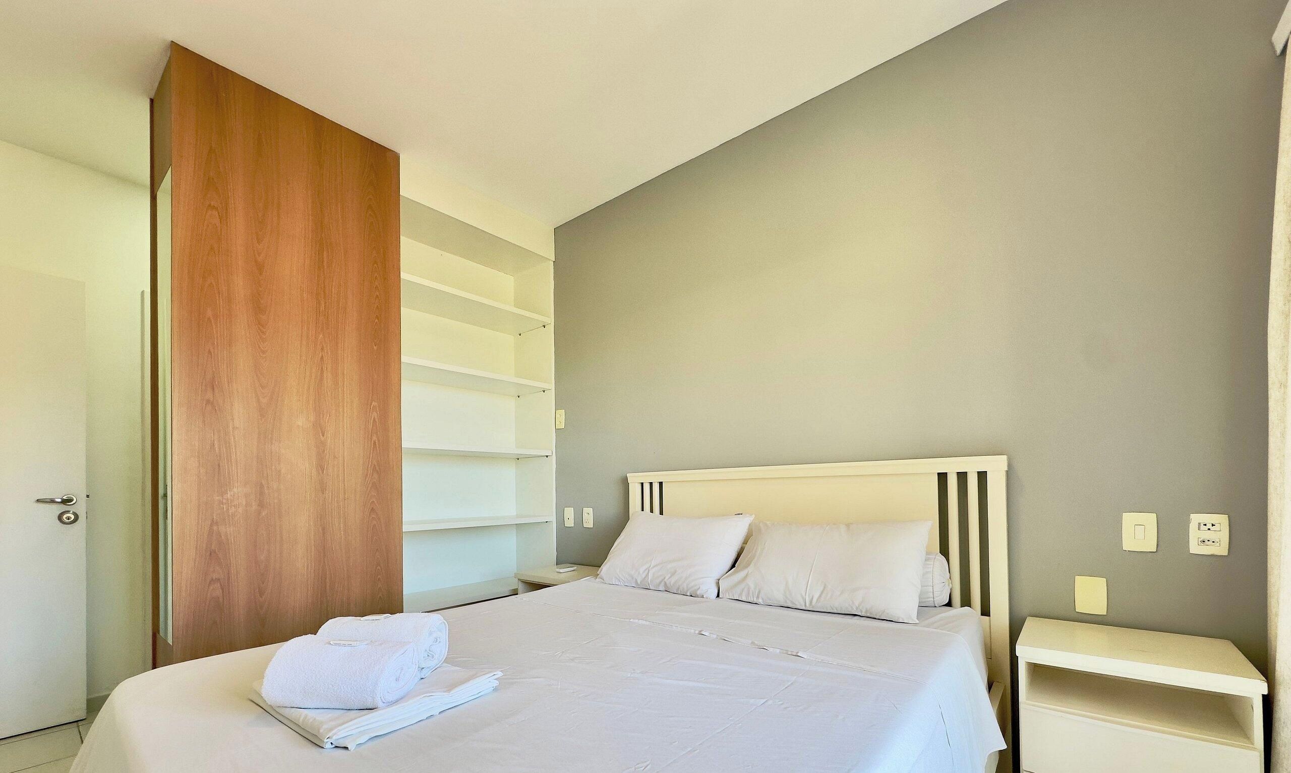 Quarto de hotel com cama, guarda-roupa de madeira e mesa de cabeceira.