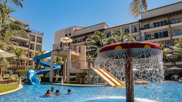Uma piscina de hotel com um tobogã, palmeiras e um edifício alto ao fundo.