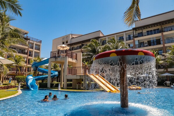 A hotel pool with a water slide, palm trees, and a multi-story building in the background.