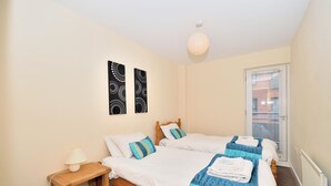 A bedroom with two beds, a bedside table, a lamp, and a door with blinds. - Bakers Court by Guestz (Chester)