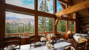 Comedor de madera con una mesa dispuesta para cuatro, grandes ventanales con vista a la montaña y una zona de estar acogedora con chimenea.