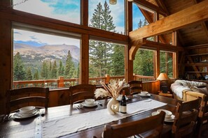 Une salle à manger en bois avec une table dressée pour quatre personnes, de grandes fenêtres offrant une vue sur les montagnes et un coin salon confortable avec une cheminée.