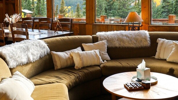 Interior de una cabaña de madera con un sofá grande, una mesa de comedor y vistas a una cadena montañosa boscosa.