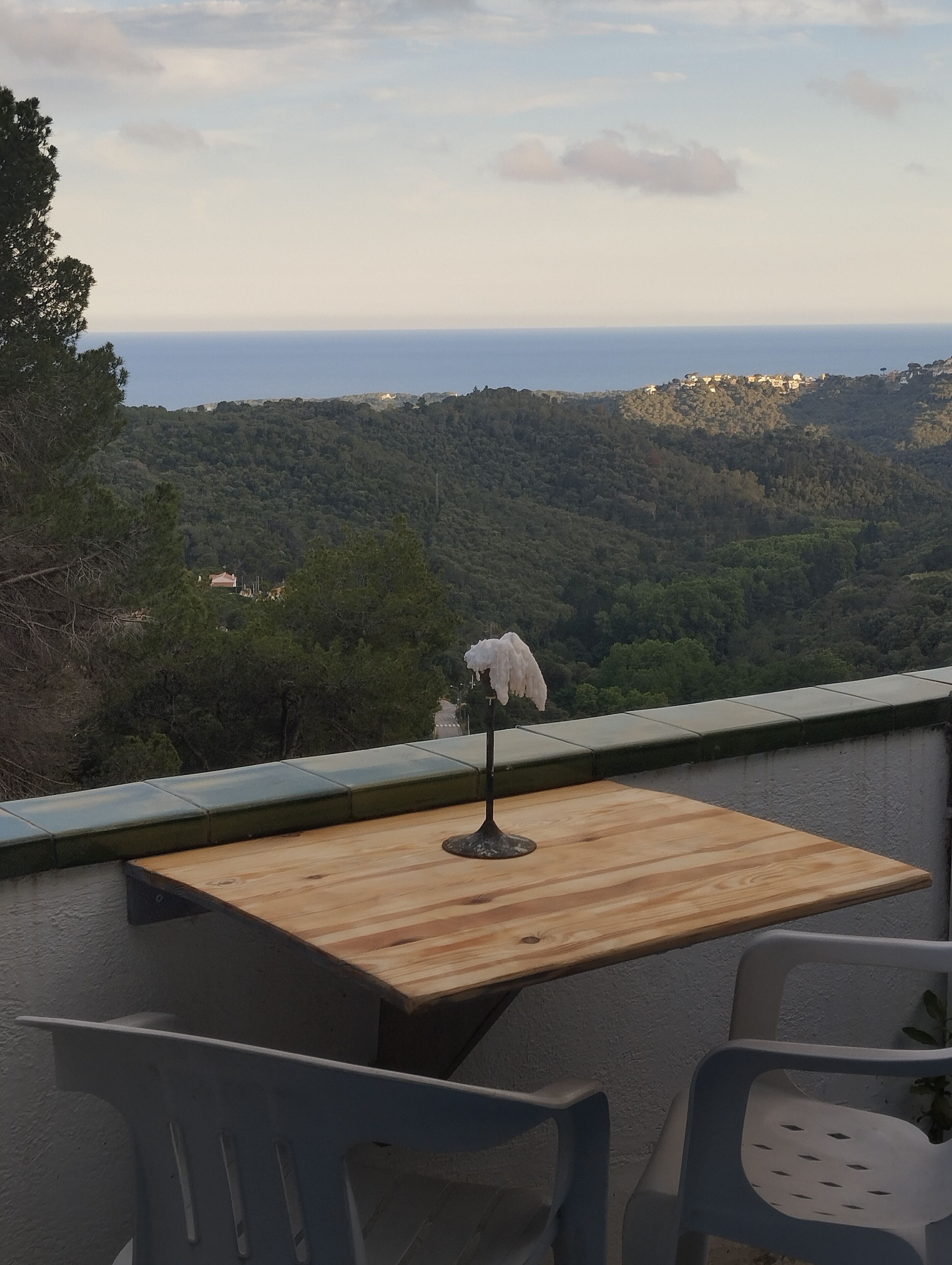 Una mesa y sillas de madera en un balcón con vistas a un paisaje boscoso y el mar.