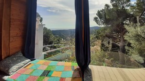 Ein Zimmer aus Holz mit Bett, einem fensterbelaubten Fenster und Blick auf Bäume und Hügel.