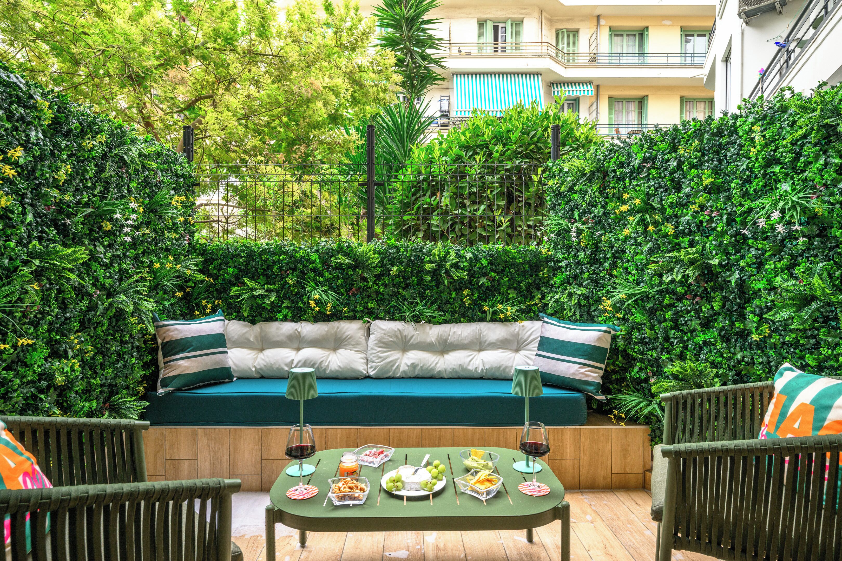 An outdoor seating area with a sofa, dining table, and chairs, surrounded by lush greenery and a building in the background.