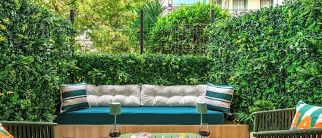 An outdoor seating area with a sofa, dining table, and chairs, surrounded by lush greenery and a building in the background.