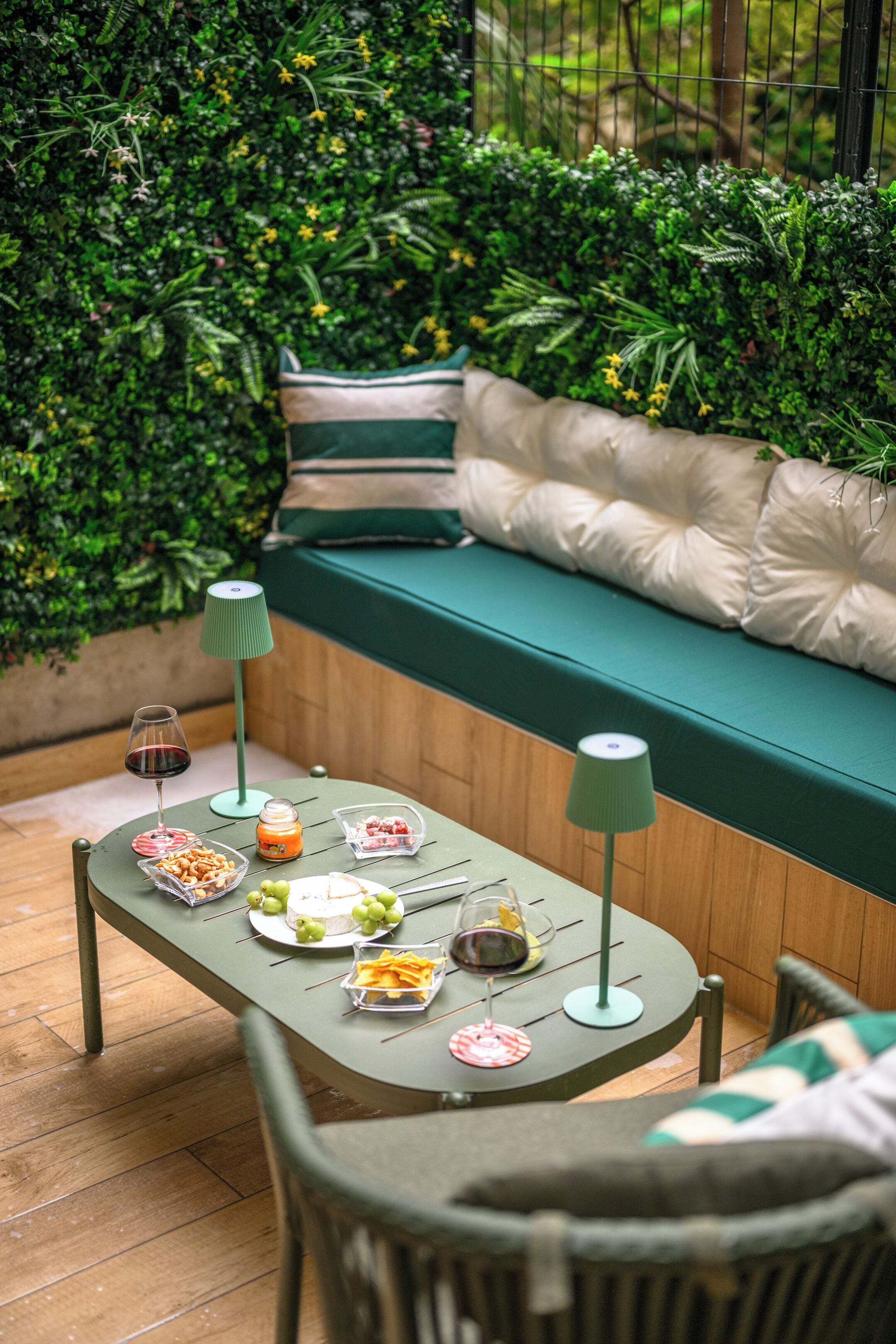A cozy outdoor seating area with a cushioned bench, a table with food and drinks, and a green backdrop of foliage.