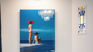 Een kamer met een blauwe bank, een schilderij van twee personen aan zee, een vaas met bloemen, en een klein tafeltje met een decoratief object.