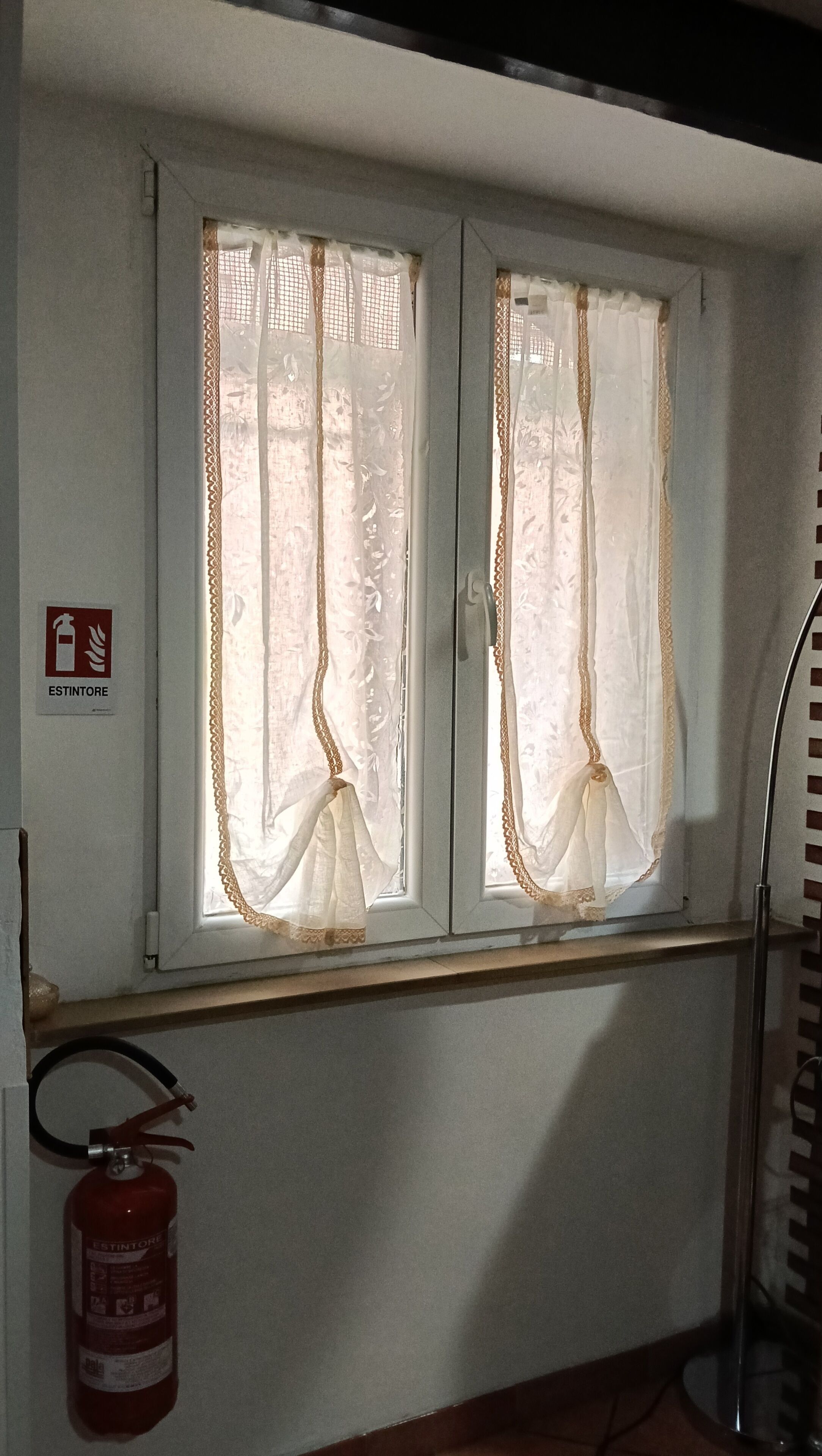 A window with sheer curtains and a fire extinguisher mounted on the wall.