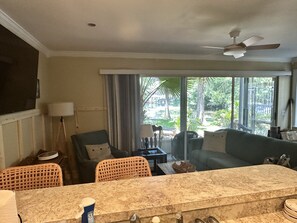 A living room with a marble coffee table, wicker chairs, a sofa, and a view of a tropical garden through large windows.