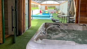 A hot tub in a covered outdoor area with a view of a swimming pool and patio furniture.