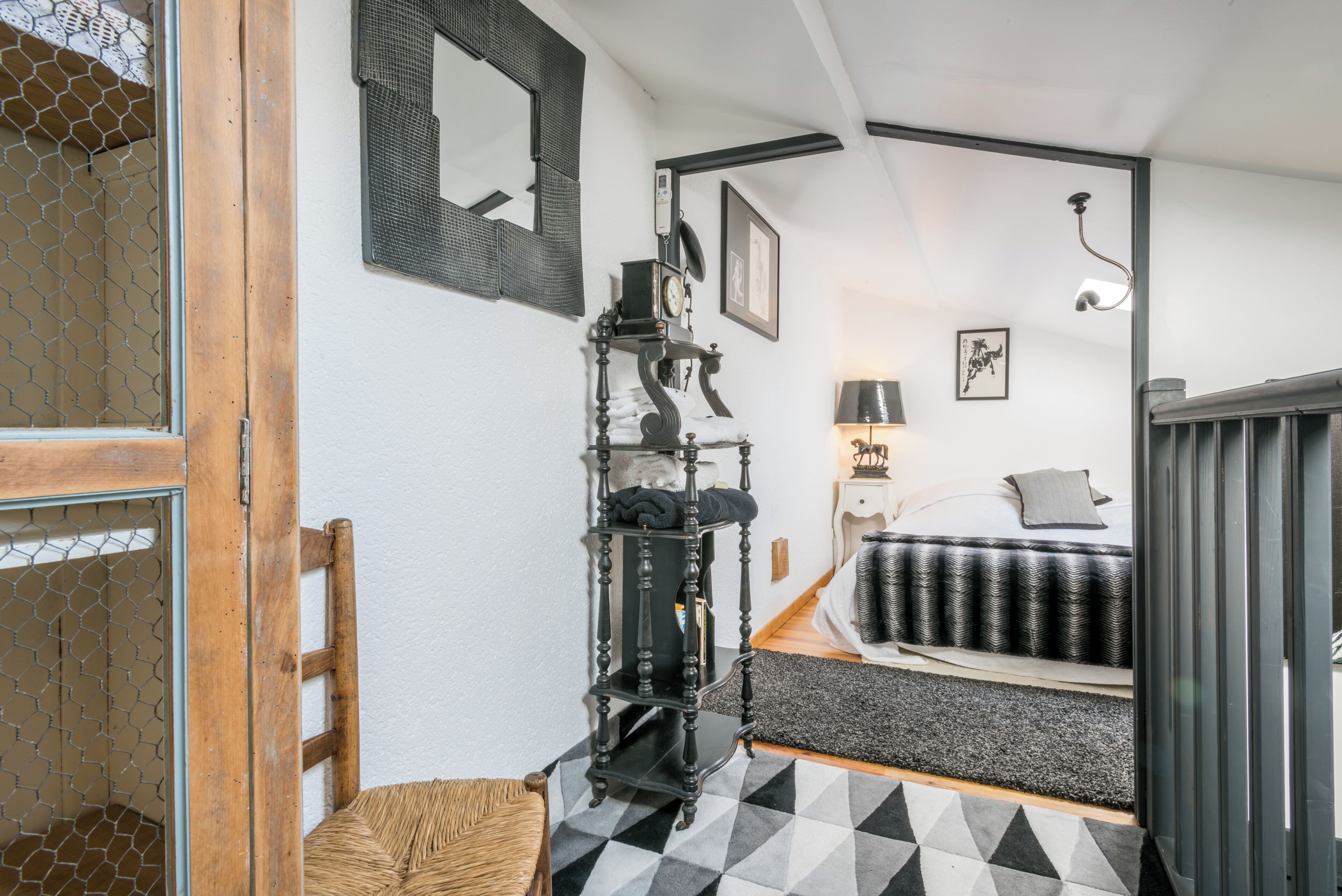 A bedroom with a bed, a bedside table, a lamp, a mirror, and a black and white patterned rug.