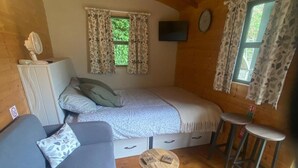 A cozy cabin interior with a bed, a sofa, a small table, and two windows with curtains. - Riverside Retreats Shepherds Hut in Newtown Wales (Newtown)