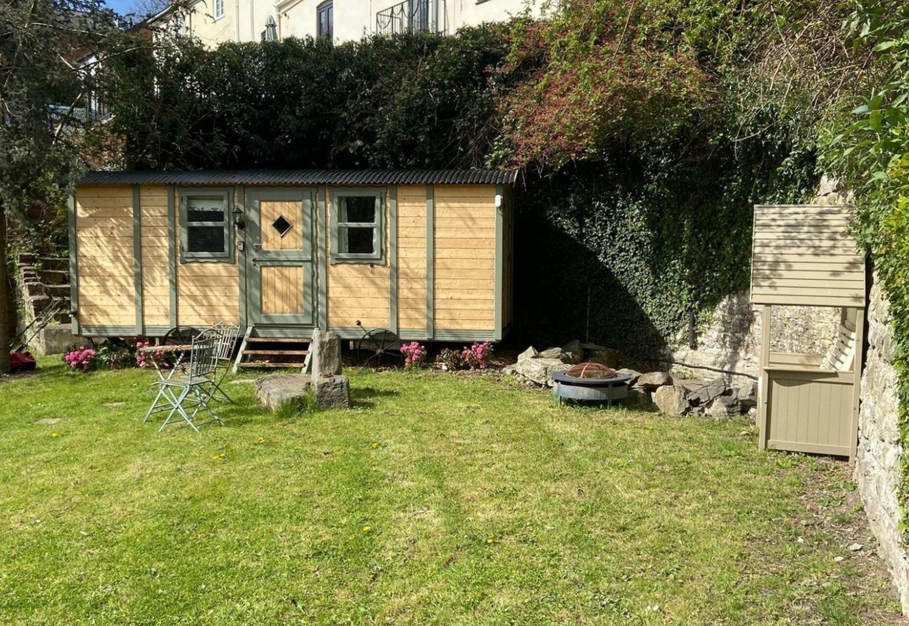 A small, single-story wooden structure with a door and windows, surrounded by a grassy yard and dense foliage.