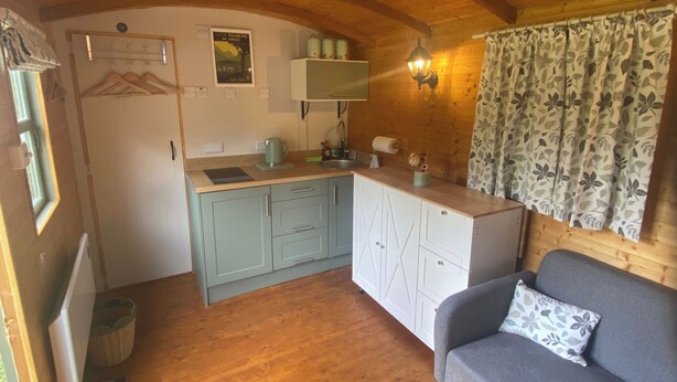 A compact kitchen with a sink, cabinets, and a small refrigerator. There is a sofa with a floral cushion and a wooden ceiling. - Riverside Retreats Shepherds Hut in Newtown Wales (Newtown)