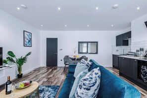 A modern living room with a blue sofa, a dining area with a small table and chairs, and a kitchen with dark cabinets and white tiles. - Binding House by Guestz (Hull)