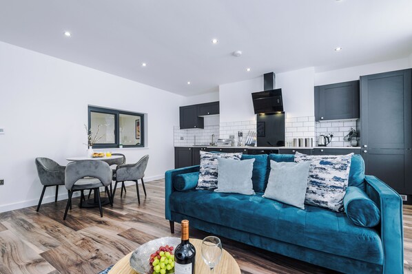 A modern kitchen with dark cabinets, a blue velvet sofa, and a dining area with grey chairs. - Binding House by Guestz (Hull)