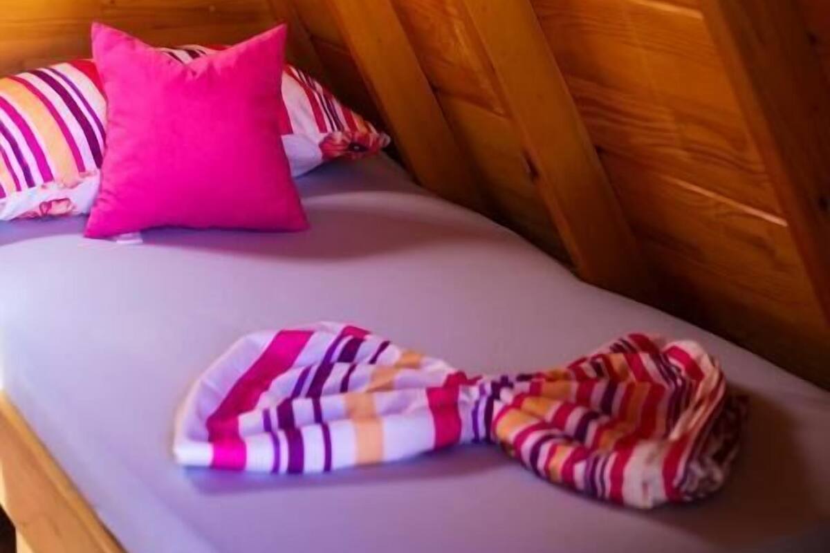A small, cozy room with wooden beams, a bed with striped bedding, and a single pink pillow.