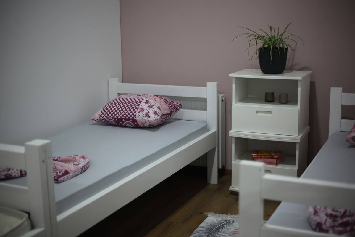 A single bed with a patterned pillow, a white bedside table, a small white nightstand, a potted plant, and a wooden floor.