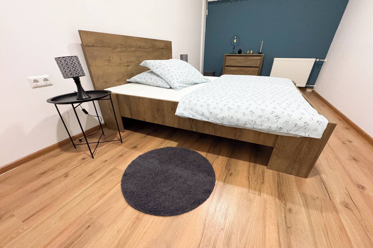 A room with a wooden bed, a small table with a lamp, a round rug, and a dresser with a mirror.
