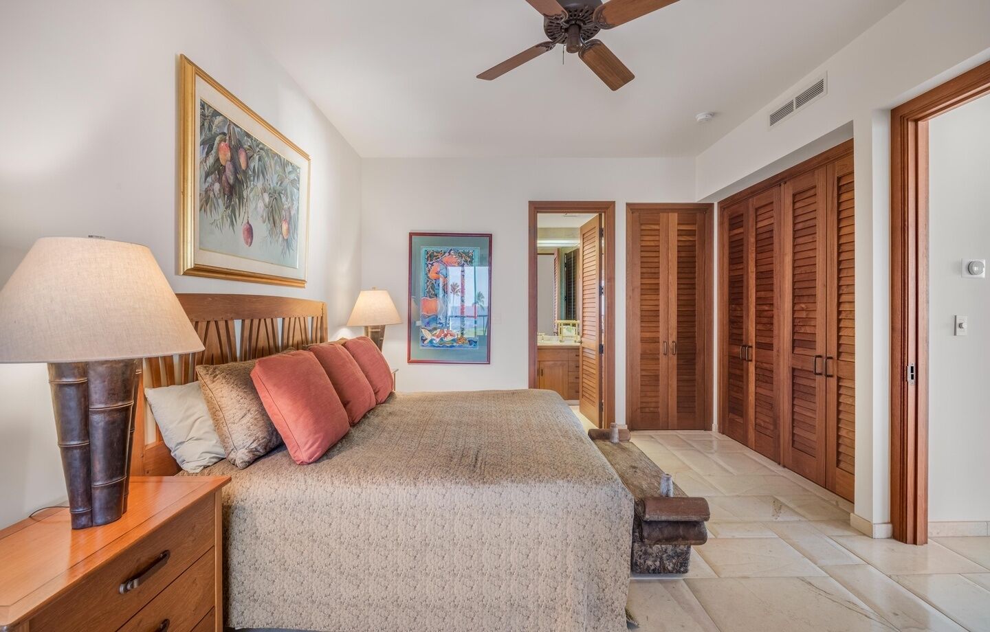 A bedroom with a bed, nightstands, a ceiling fan, wooden doors, and artwork on the walls.