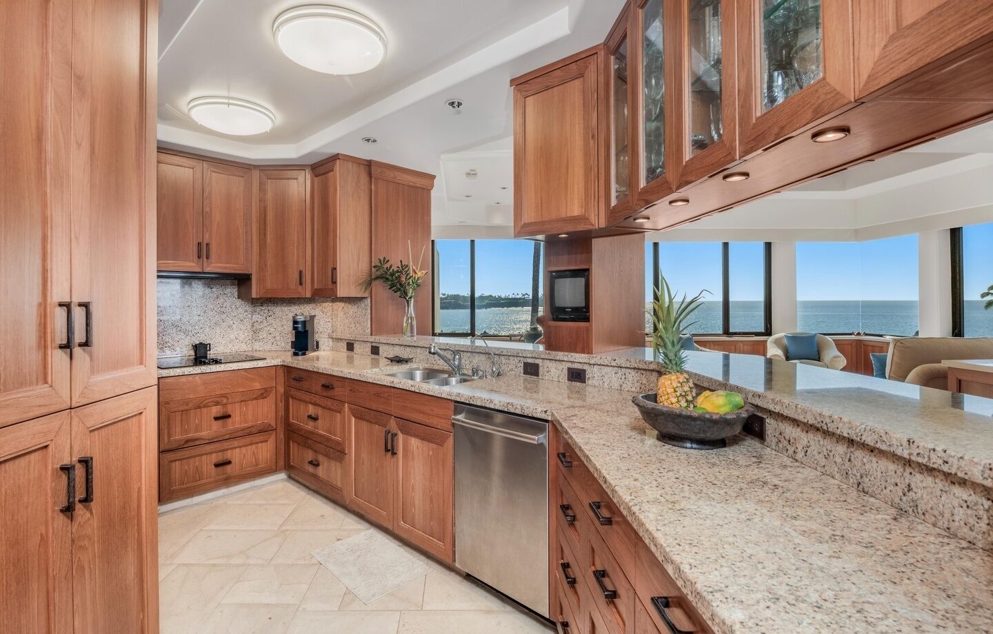 A modern kitchen with wooden cabinets, granite countertops, a stainless steel dishwasher, and a large window offering a view of the ocean.
