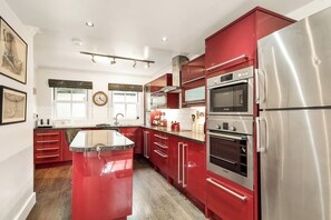 A kitchen with red cabinets, stainless steel appliances, a central island, and a window with a view. - Spacious 3BR Home w/ Private Garden & Parking (Cambridge)