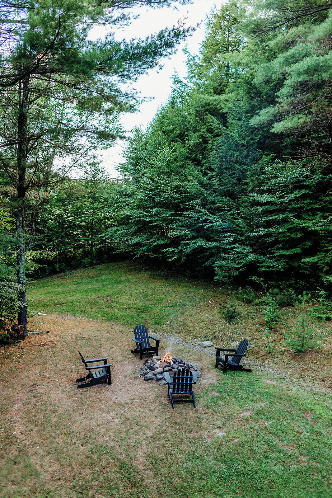 A campsite with three Adirondack chairs arranged around a fire pit in a wooded area.