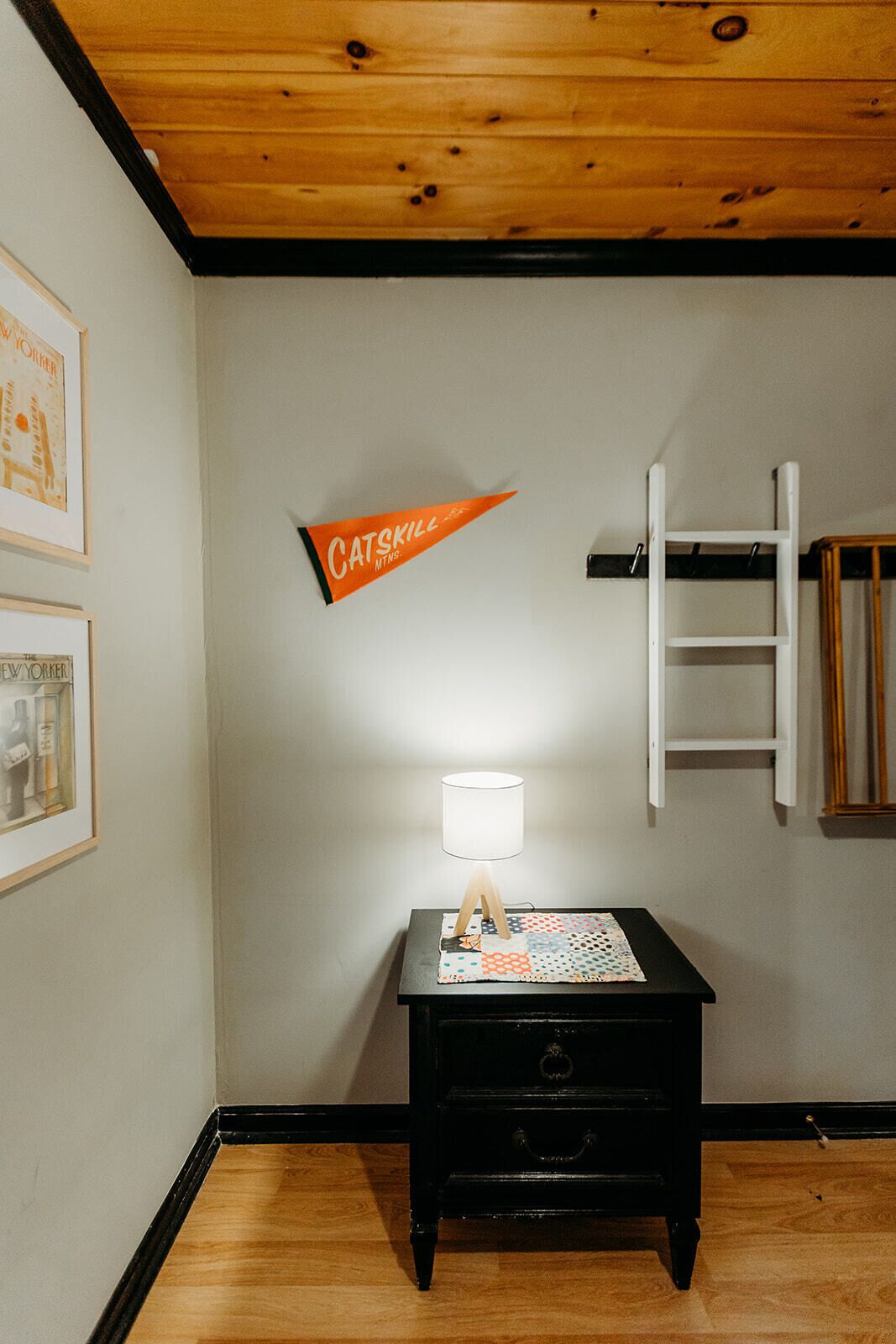 Een kamer met een houten plafond, een zwart nachtkastje met lamp en een aan de muur bevestigde ladder, plus een lijst met een afbeelding.