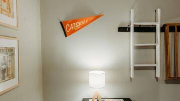 Een kamer met een houten plafond, een zwart nachtkastje met lamp en een aan de muur bevestigde ladder, plus een lijst met een afbeelding.