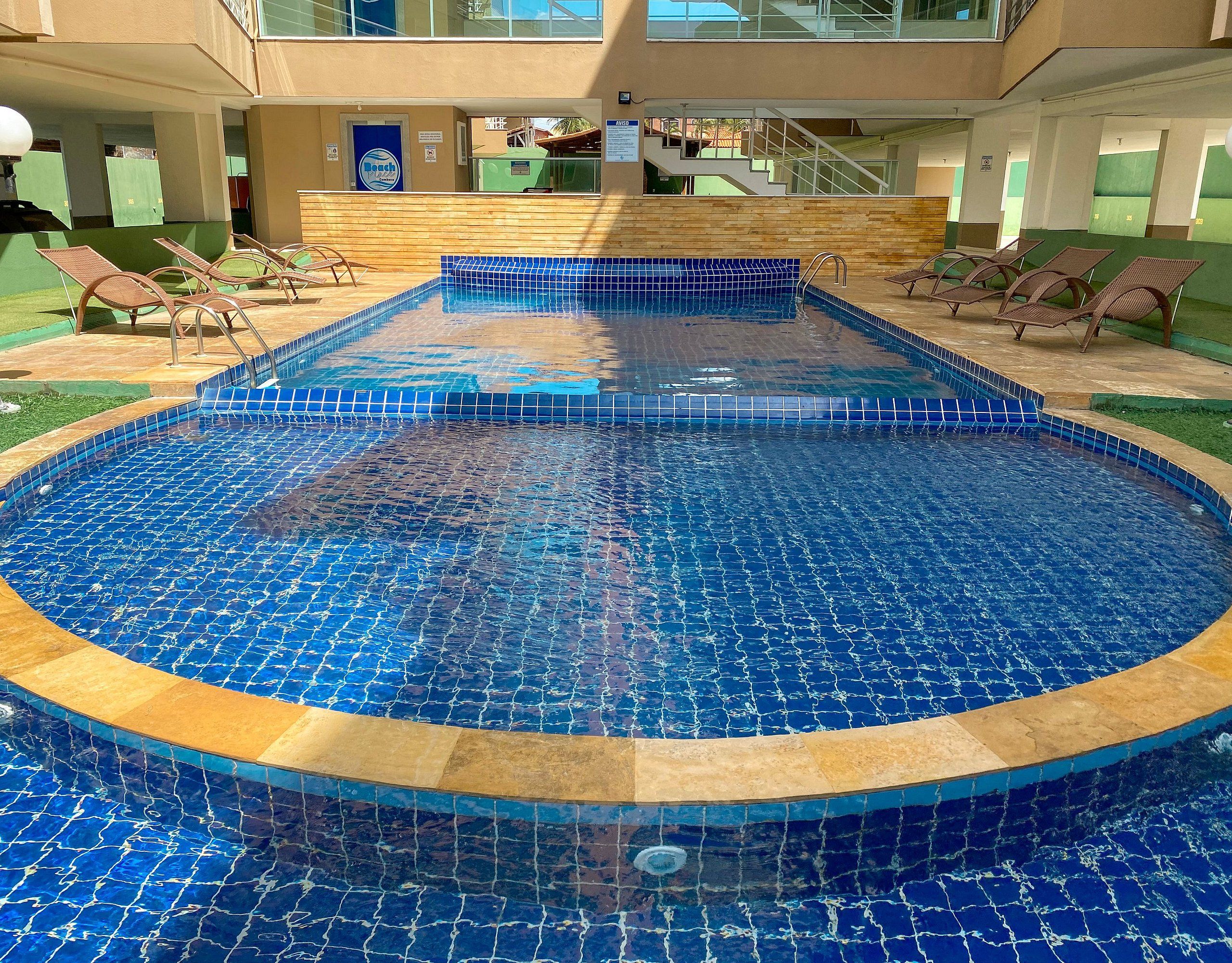 Piscina com borda azulejada, rodeada por espreguiçadeiras e um edifício com varanda.