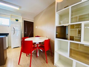 A compact kitchen with a white refrigerator, a dining table with red chairs, and a white shelving unit with glass doors. - Lindo Apto no Cumbuco por Be My Guest (Caucaia)