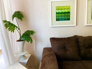 Un salon avec un canapé marron, une table d’appoint blanche, une plante en pot et un tableau abstrait encadré accroché au mur.