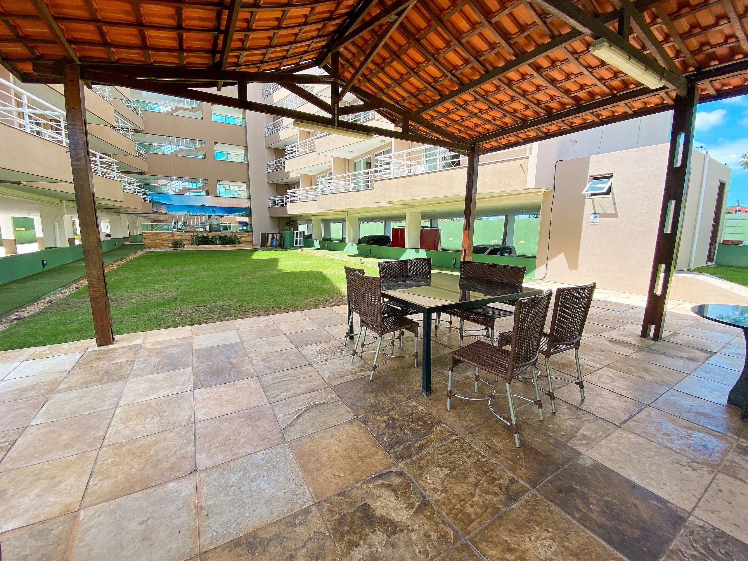 A covered outdoor area with a table and chairs, a grassy lawn, and a multi-story building in the background.